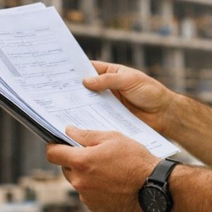 Profissional de obras usando capacete e colete refletivo observa atentamente um edifício em construção enquanto segura e confere documentos nas mãos