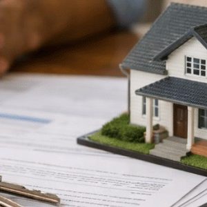 Documentos espalhados sobre uma mesa de madeira, com uma maquete de casa e um conjunto de chaves posicionados sobre os papéis, sugerindo um contexto de compra, venda ou regularização de imóvel. A cena tem iluminação natural suave e fundo levemente desfocado, transmitindo um ambiente institucional ou administrativo.